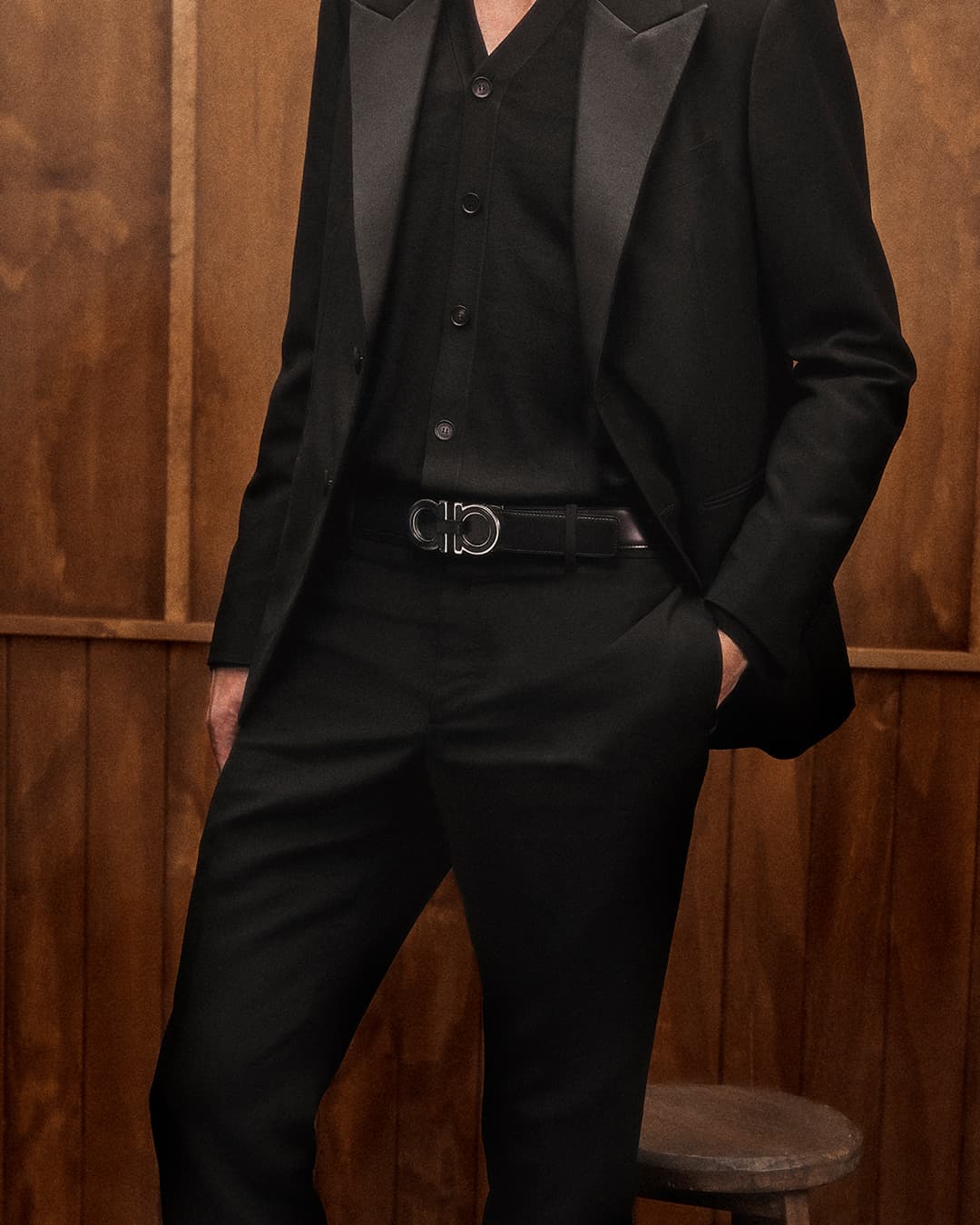 Fashion shot of a man in a black suit with a black button-down shirt and a belt with a silver buckle against a wood background.
