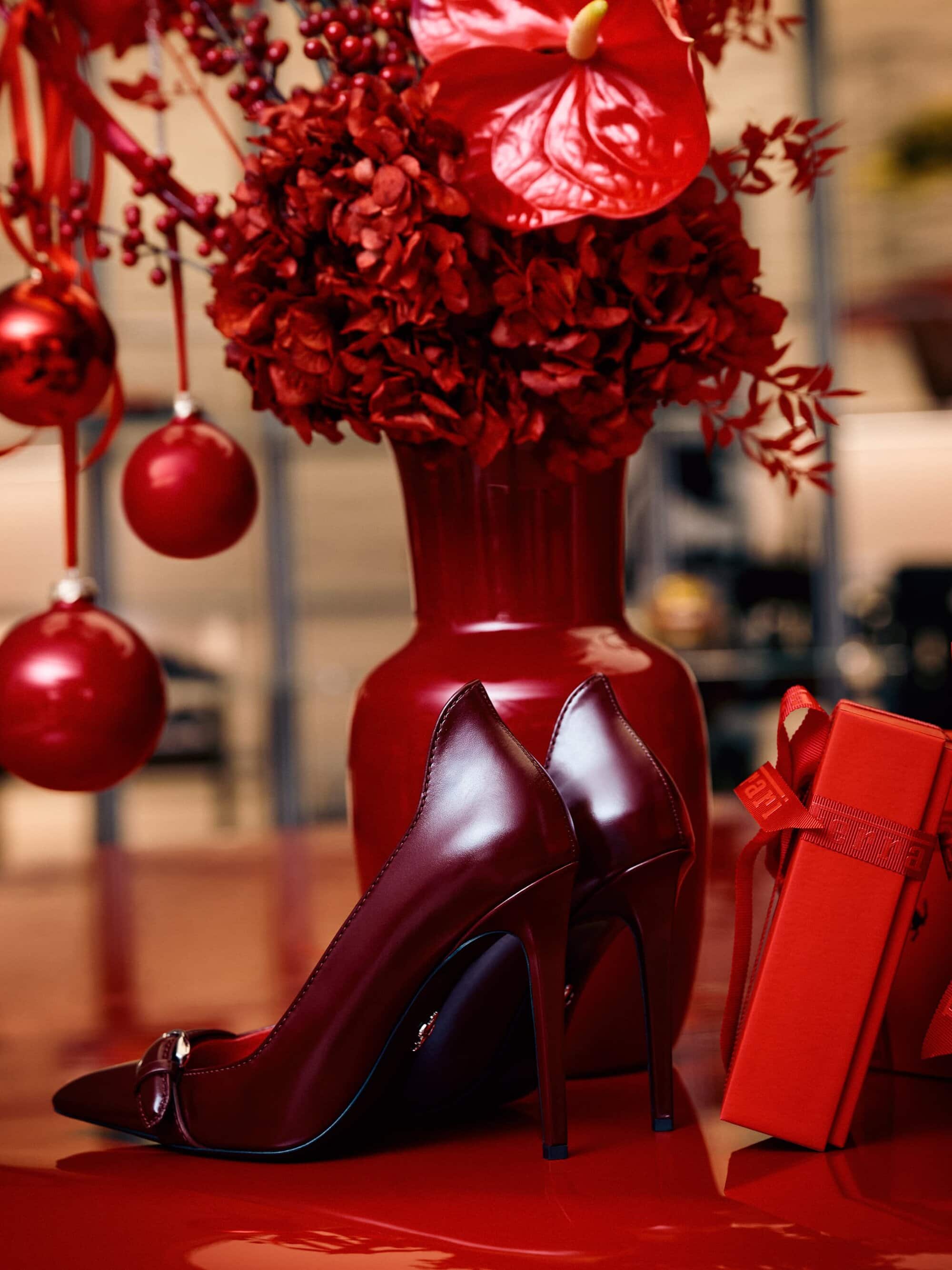 Burgundy pumps on a glossy surface next to a red vase with flowers, Christmas ornaments, and a gift box, all in a studio.