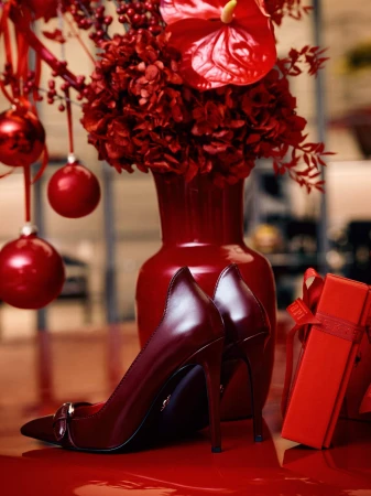 Burgundy pumps on a glossy surface next to a red vase with flowers, Christmas ornaments, and a gift box, all in a studio.