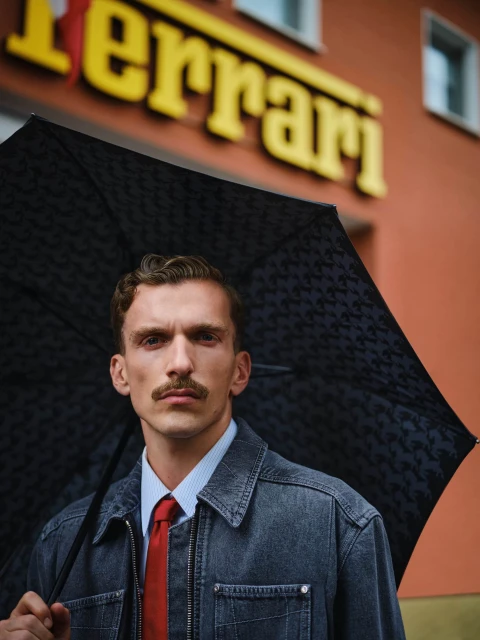 Adult man with a mustache posing with an umbrella in front of a building with Ferrari signage in an outdoor, medium shot.