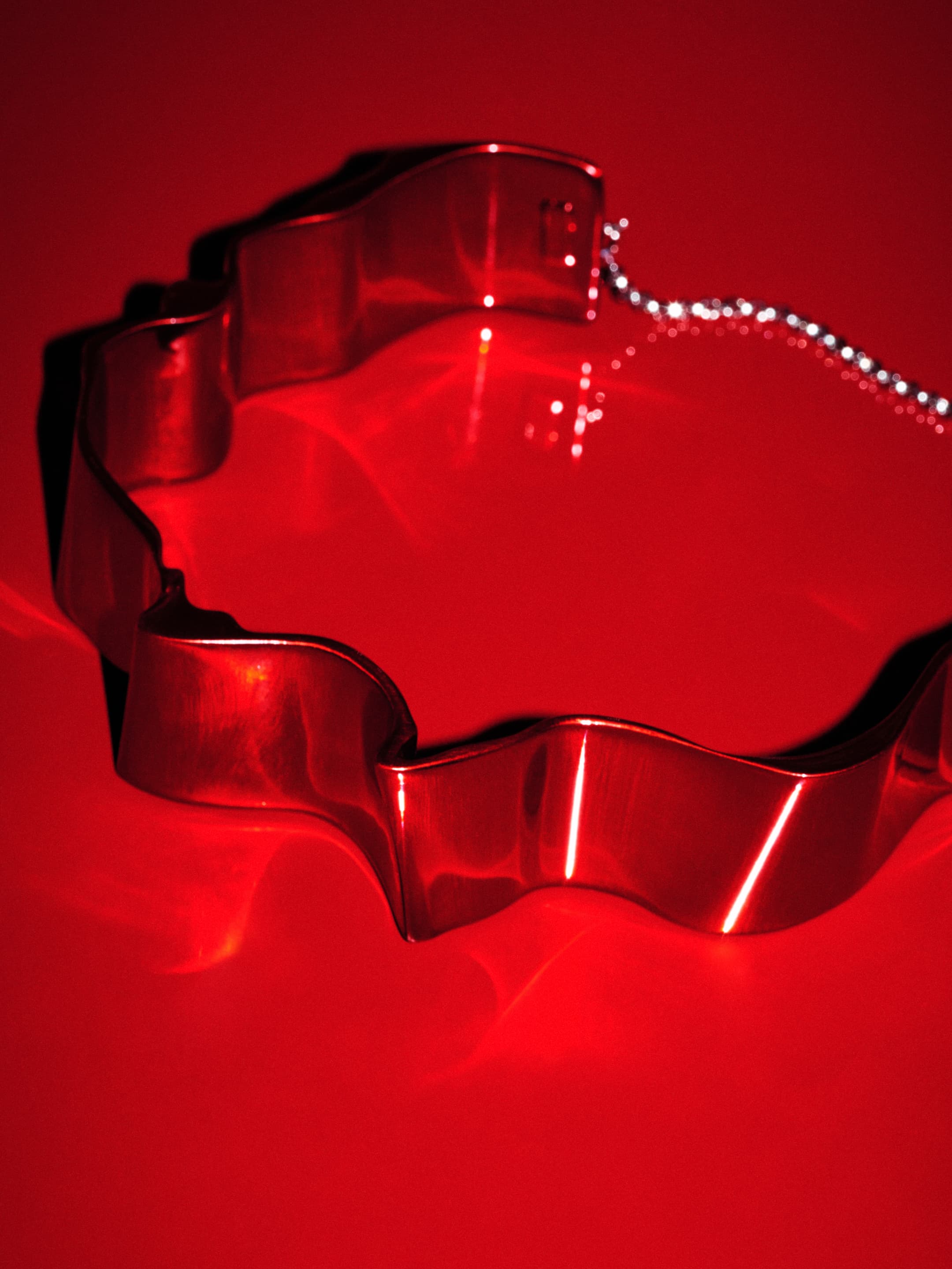 Shiny metal wavy cookie cutter with silver chain detail lies on a seamless background and reflects bright red lighting in a studio shot.