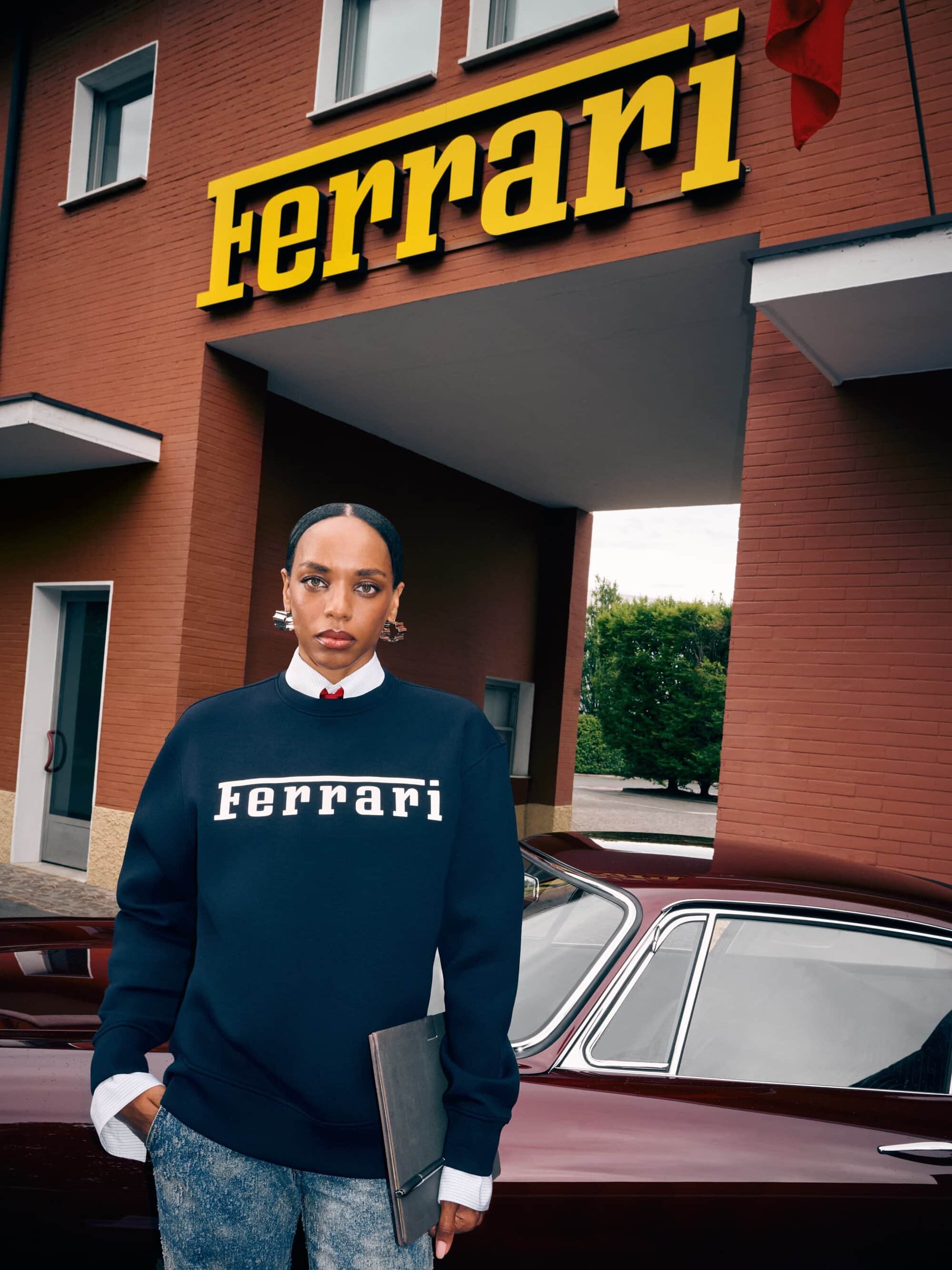 Woman in Ferrari sweatshirt standing by a maroon car, wearing silver earrings, grey pants, and holding a grey folder in front of brick building.