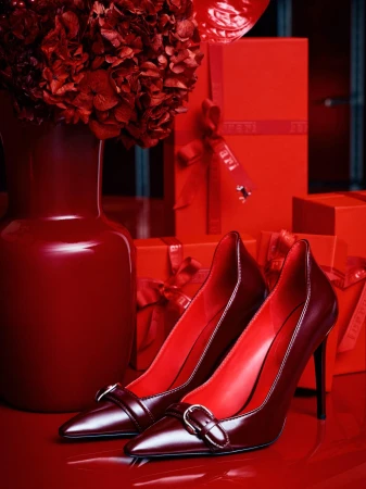 A pair of red high-heel shoes sit on a glossy surface next to a red flower arrangement and red gift boxes in a still life.