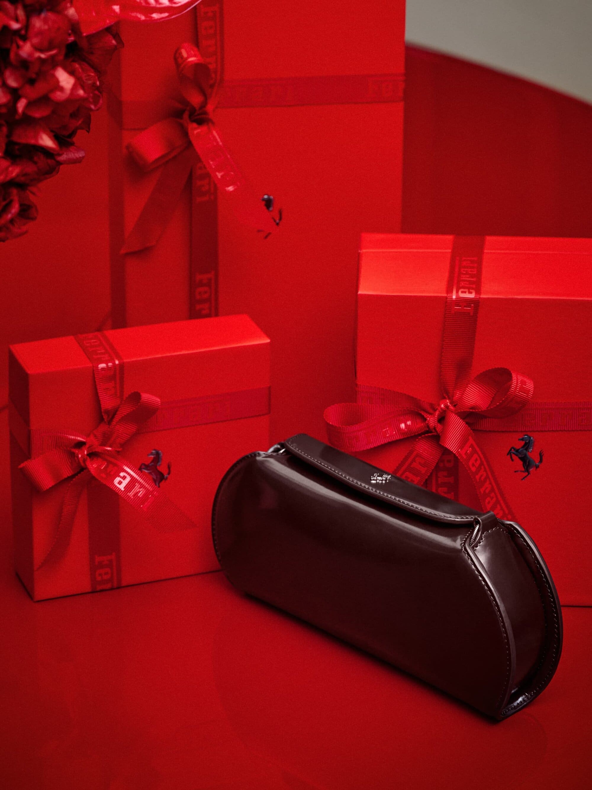 Display of gift-wrapped red boxes with red ribbons and a dark brown leather clutch on a glossy red surface, with flowers in background.