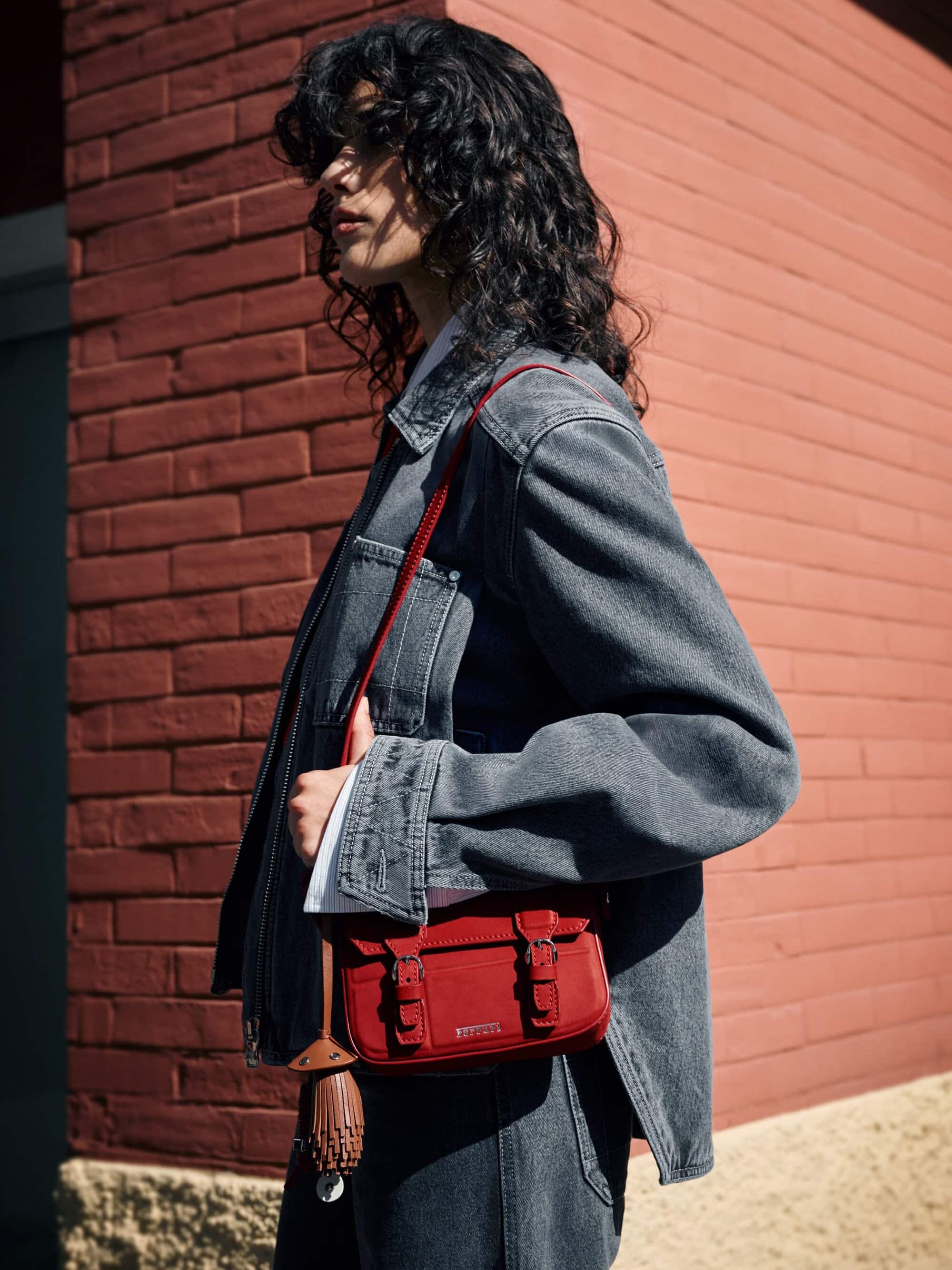 A woman stands in front of a brick wall wearing a denim jacket and jeans, holding a red satchel purse with a brown umbrella. 