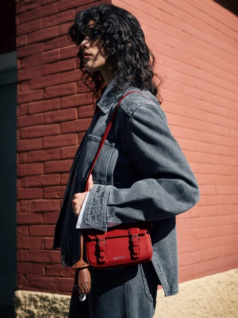 A woman stands in front of a brick wall wearing a denim jacket and jeans, holding a red satchel purse with a brown umbrella. 