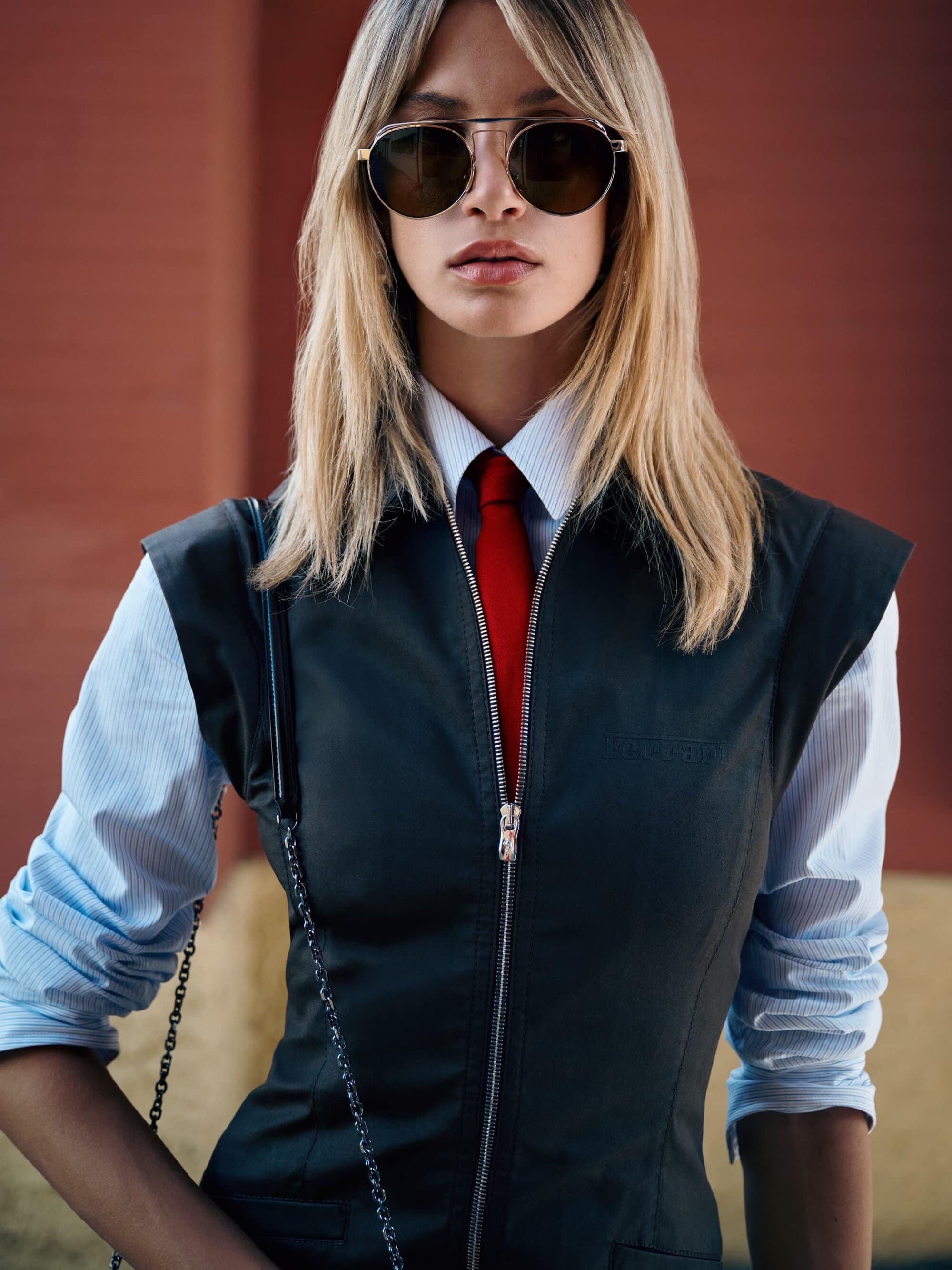A striking fashion shot of a blonde woman in aviator sunglasses, a blue striped shirt, red tie, and black dress with silver zipper.