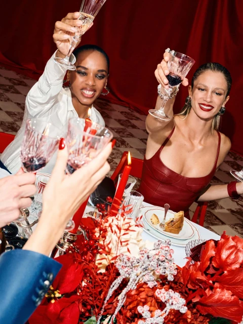 Two women toasting with wine glasses at a holiday dinner. The table is decorated with red flowers, berries and candle, set against a red backdrop.