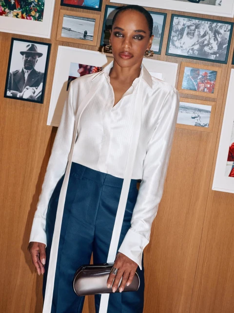 Elegant woman in white blouse and blue pants with white stripes, holding a metallic clutch, posing in front of framed photos.