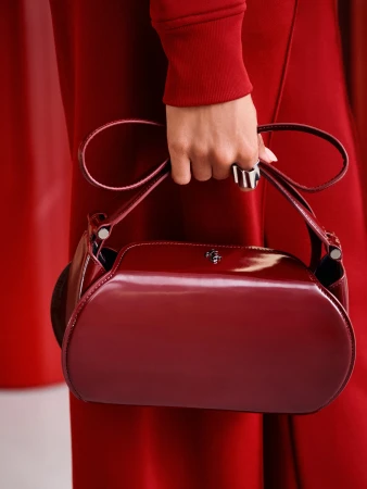 Close-up of an adult female holding a shiny, dark red handbag against a matching monochromatic background, showcasing fashion accessory.