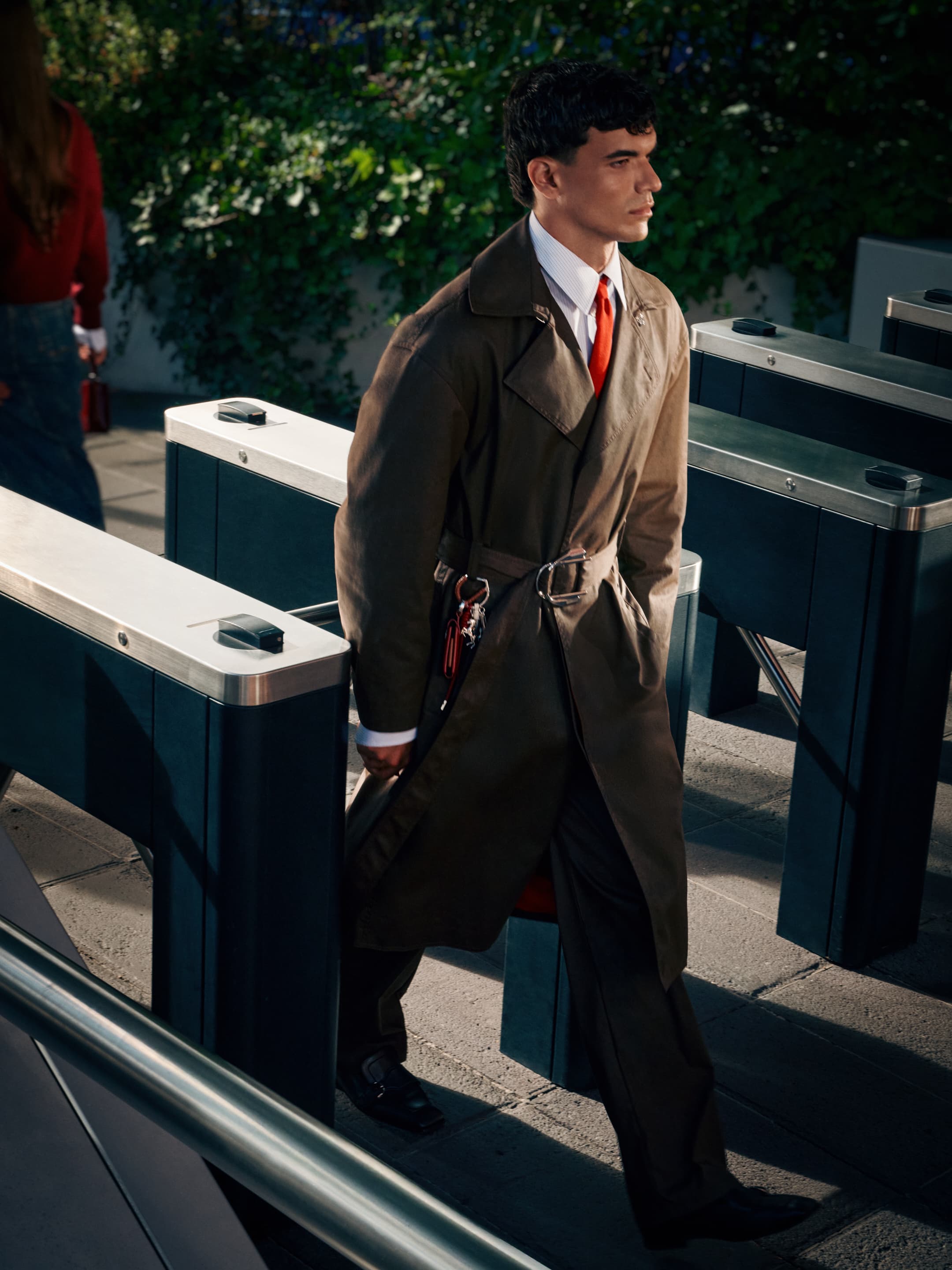 Adult male in brown trench coat walks through turnstile in an outdoor urban setting with greenery and pedestrian traffic in the background.