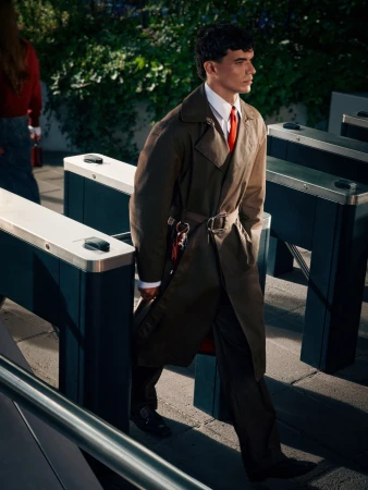 Adult male in brown trench coat walks through turnstile in an outdoor urban setting with greenery and pedestrian traffic in the background.