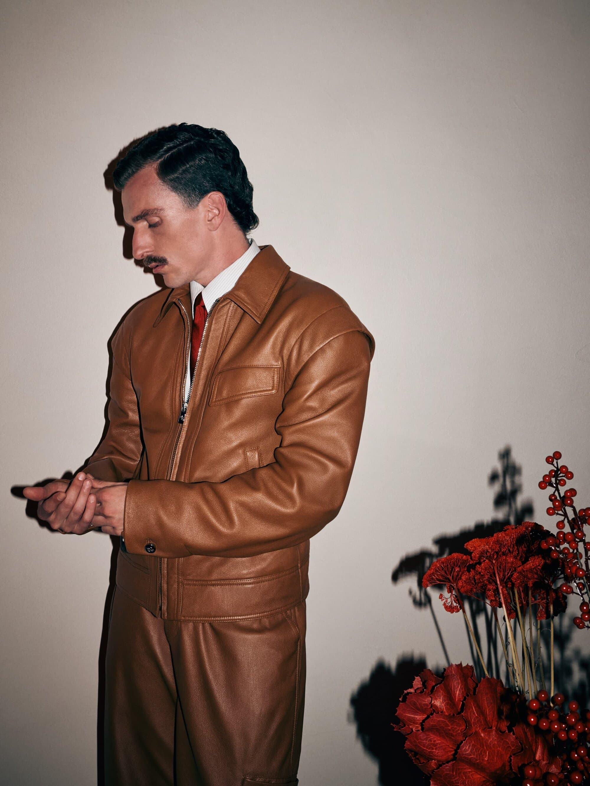 Adult male in a brown leather jacket and pants standing indoors near a red flower arrangement, showcasing a blend of fashion and style.
