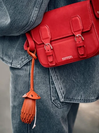 Close-up of a red Ferrari handbag with a tassel attached, against a blue denim jacket, creating a high-contrast fashion accessory shot.