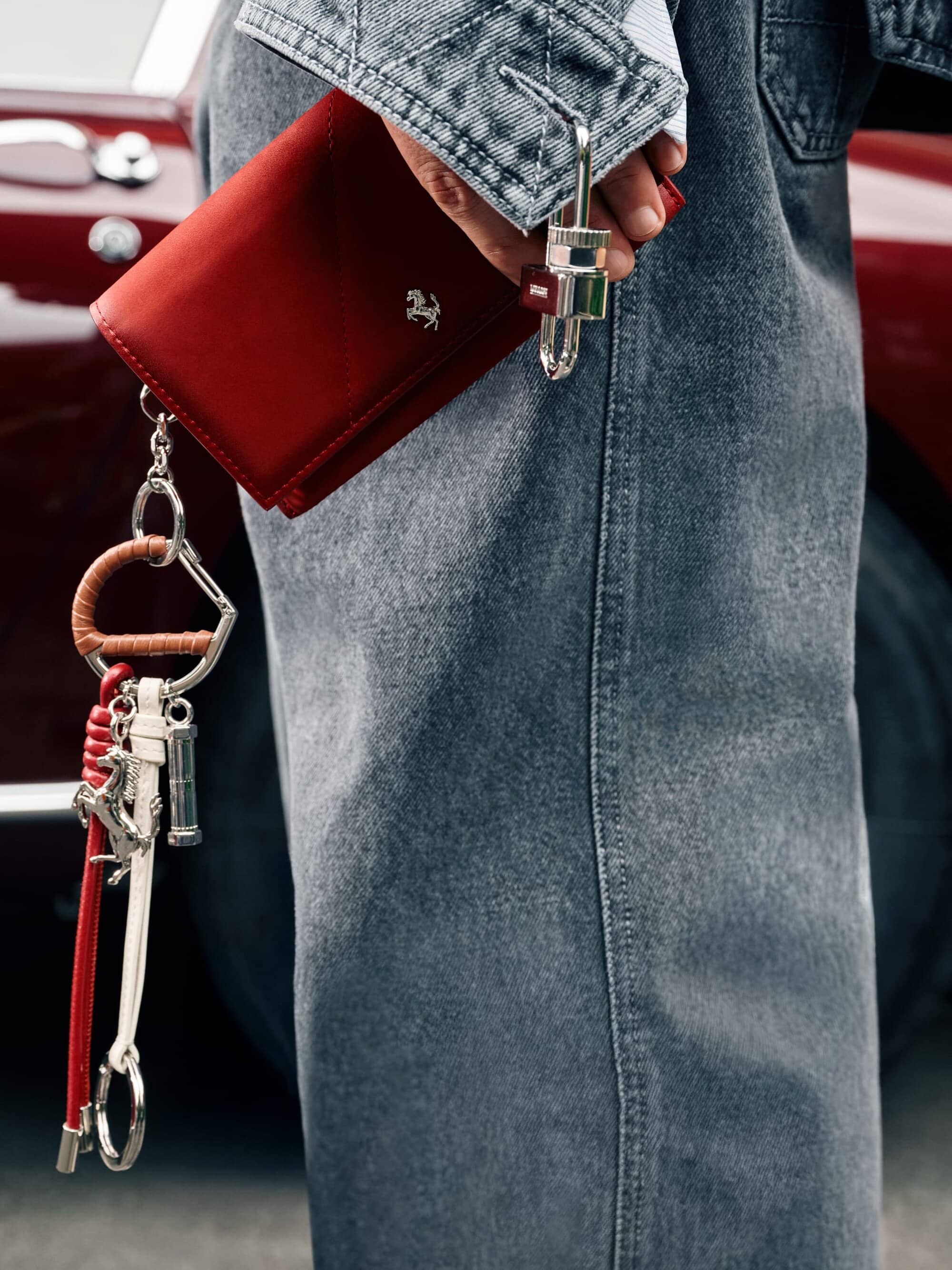 Close-up of a person holding a red leather wallet with a silver keychain attachment, set against a red car and a denim jacket.