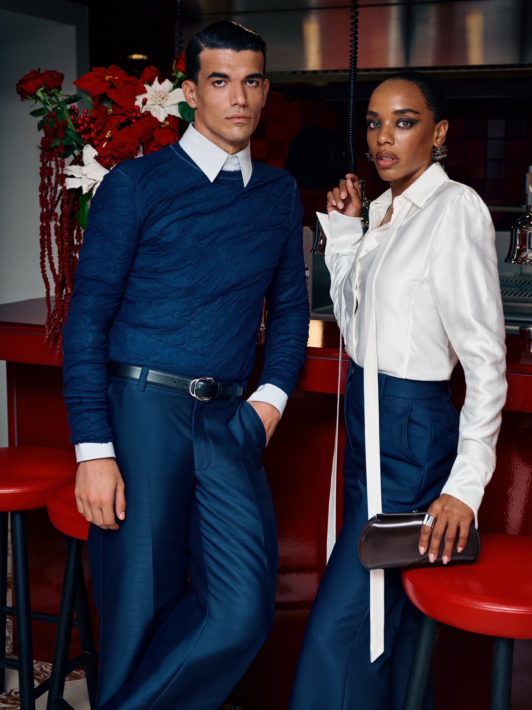 Two fashionably dressed adults, a man and woman, pose in a bar with a red counter, exuding a stylish, coordinated look for a fashion editorial.