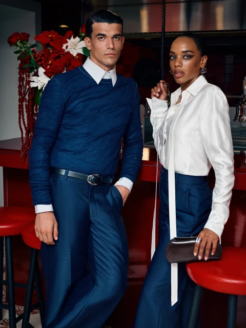 Two fashionably dressed adults, a man and woman, pose in a bar with a red counter, exuding a stylish, coordinated look for a fashion editorial.