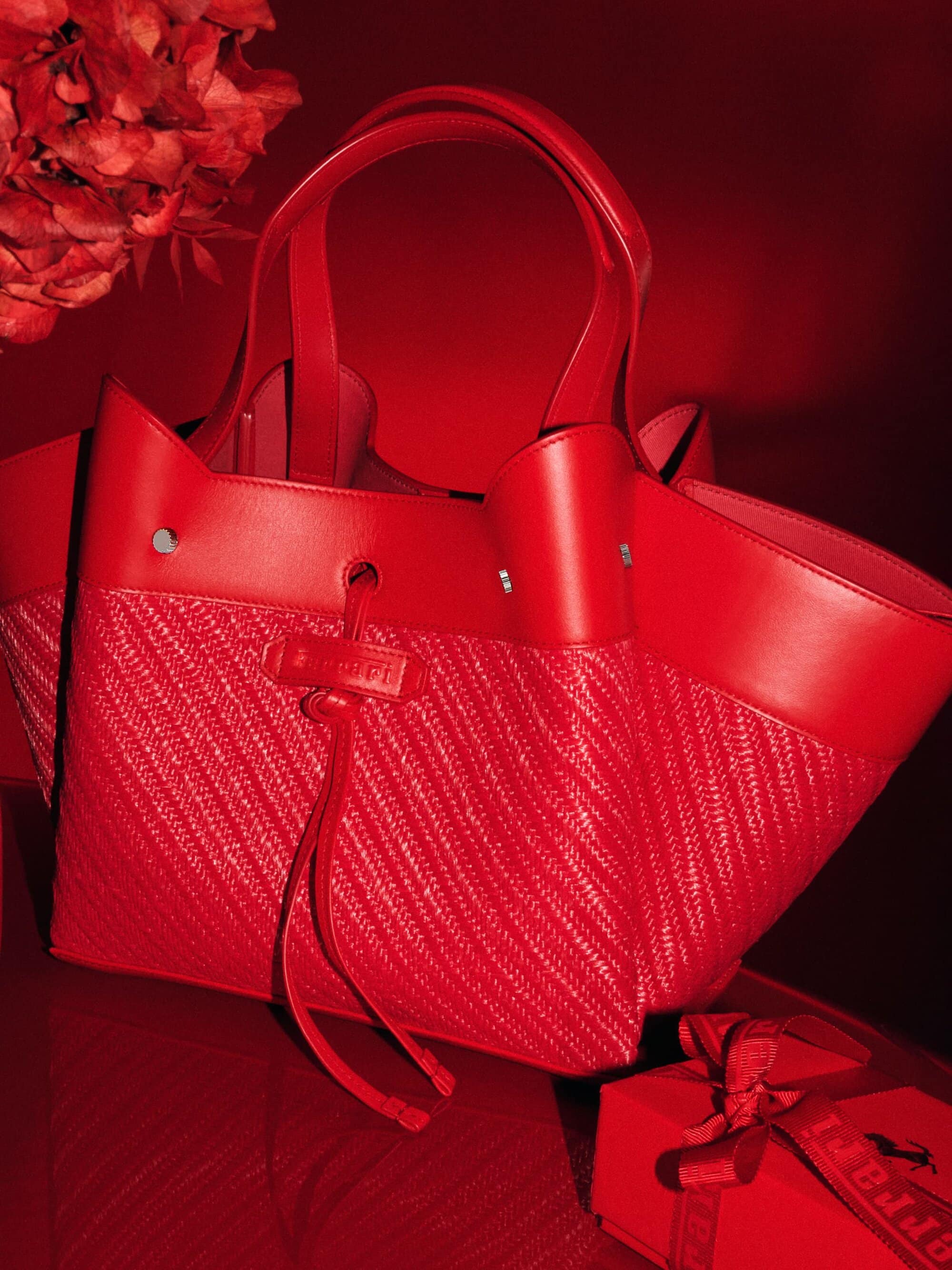 Close-up of a red leather and straw handbag with a small red gift box, all under a dramatic red light. Fashion accessory product shot.