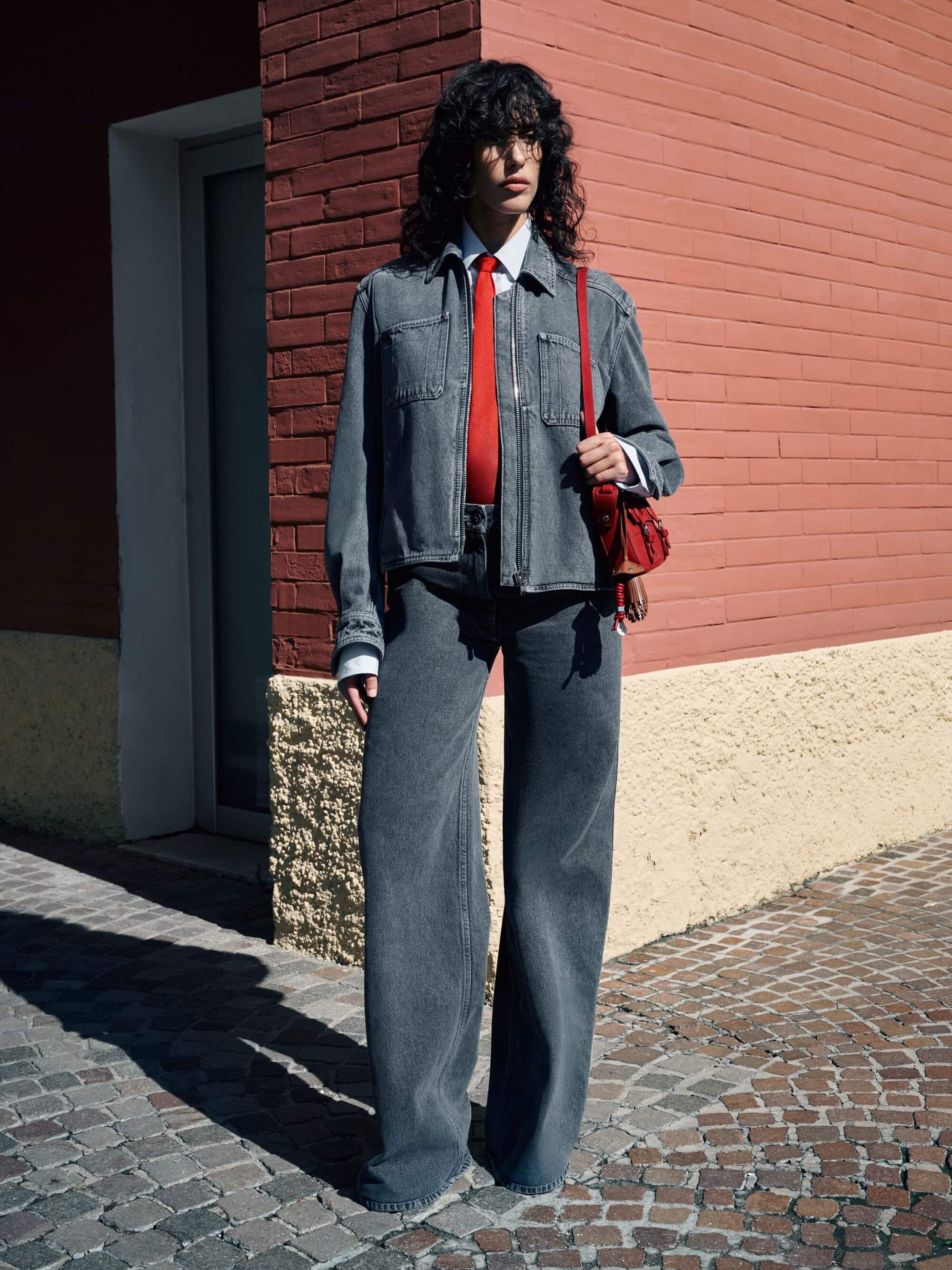 Adult female wearing wide-leg denim jeans, denim jacket and red tie posing in front of a brick building on a cobblestone street.