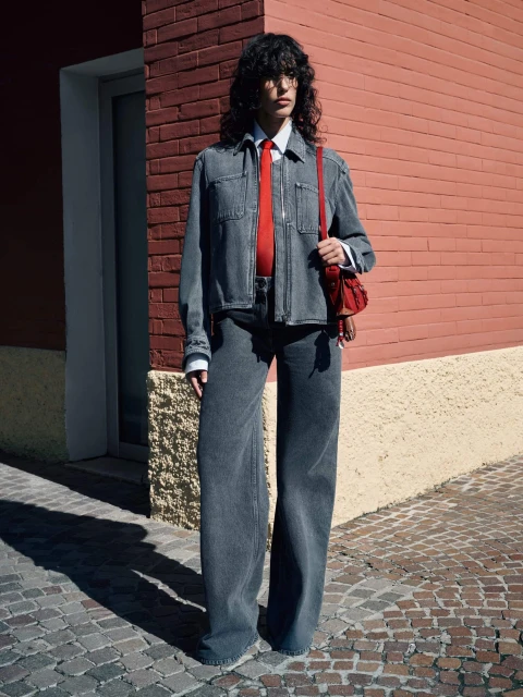Adult female wearing wide-leg denim jeans, denim jacket and red tie posing in front of a brick building on a cobblestone street.