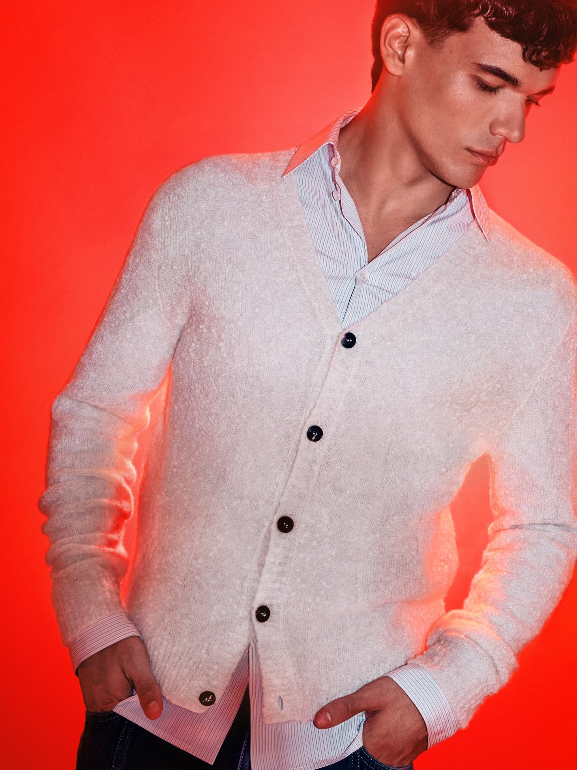 A young adult man poses against a vibrant red background, wearing a light-colored cardigan over a blue striped shirt and jeans.