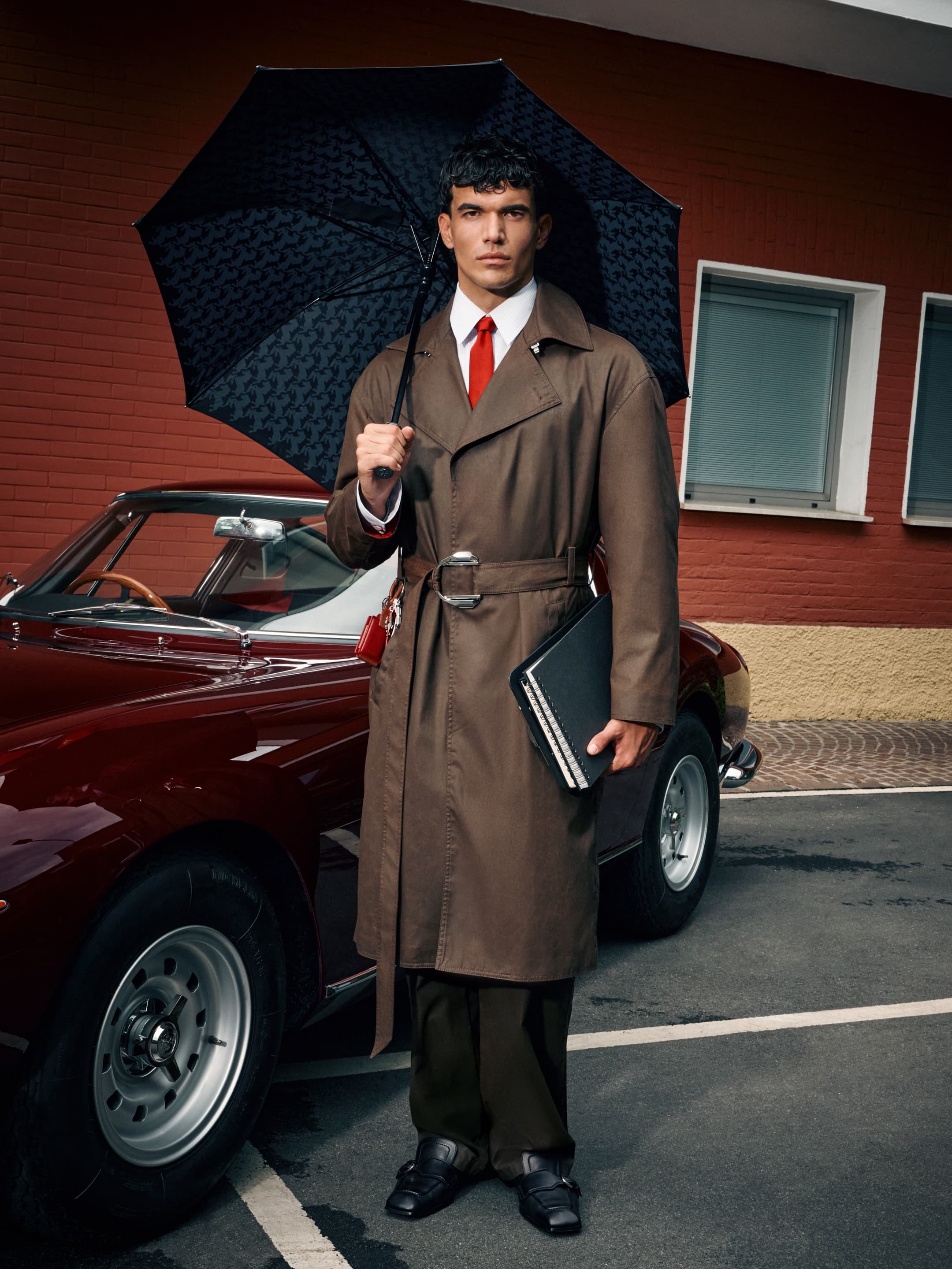 A man stands beside a car holding an umbrella and notepad wearing a brown trench coat, red tie, and dark pants on a city street.