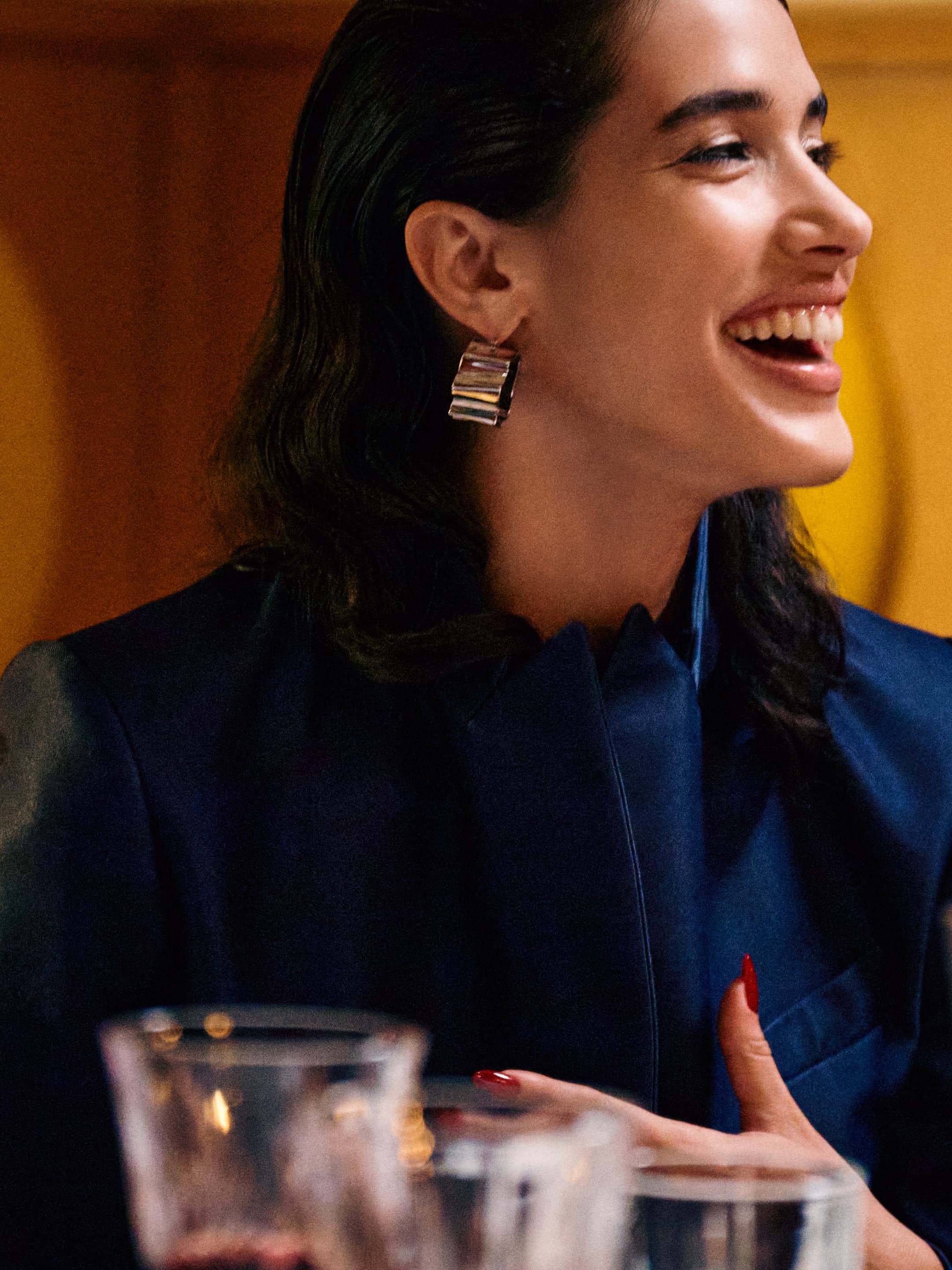 Close-up shot of a smiling woman with dark hair wearing statement earrings and a blue jacket, set indoors with glasses on the table.