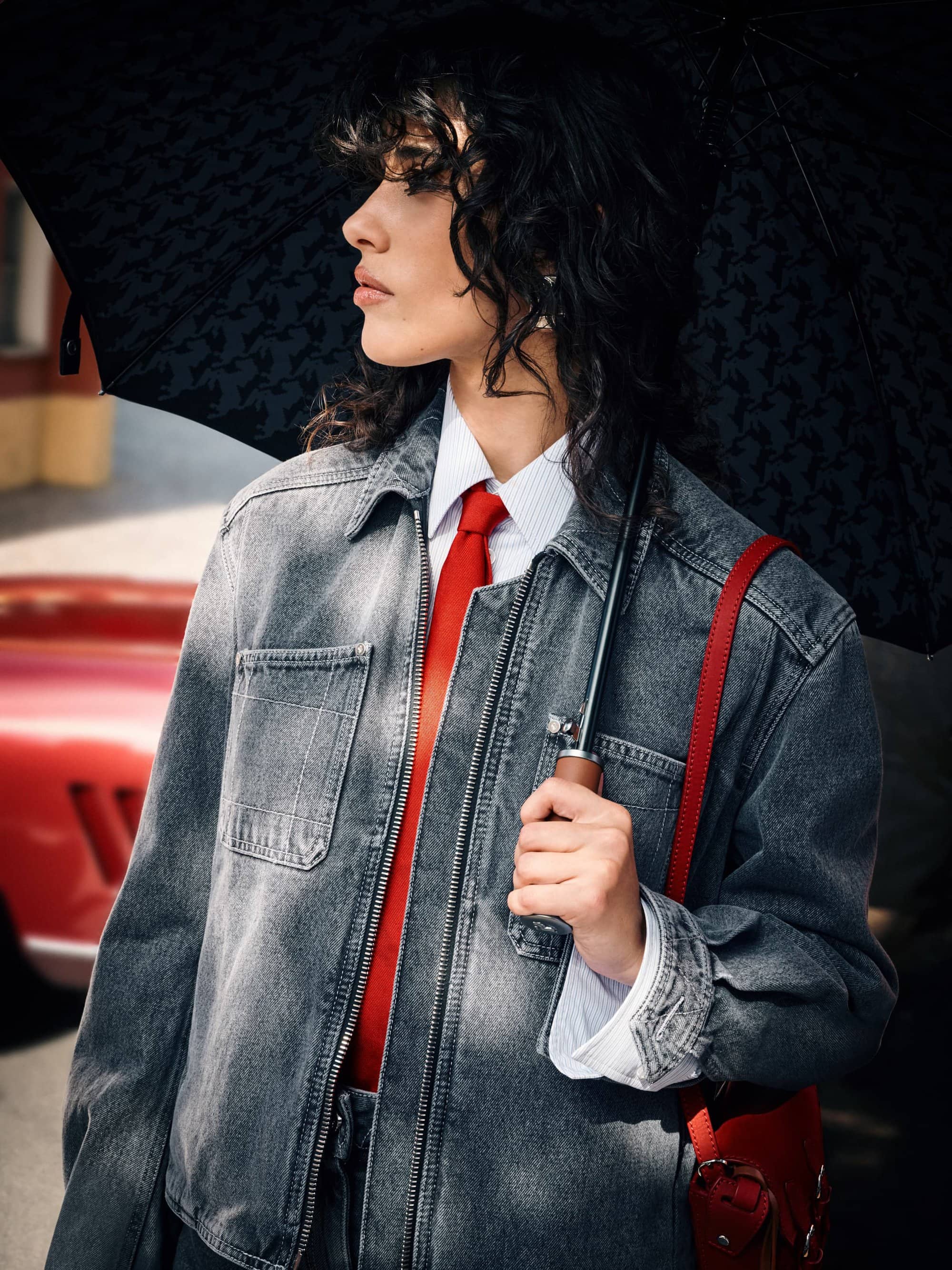 Portrait of a woman with curly hair wearing a denim jacket, a red tie, and holding a black umbrella with a red car in the background.