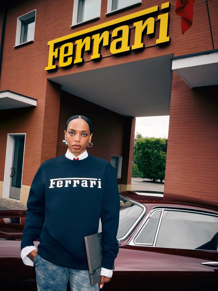 Woman wearing a dark sweatshirt with Ferrari branding posing in front of a classic car and brick building exterior.