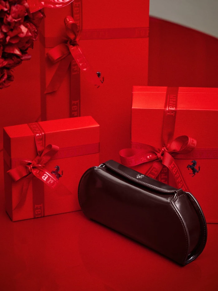 Dark glossy handbag placed in front of several stacked red gift boxes with ribbons against a red background