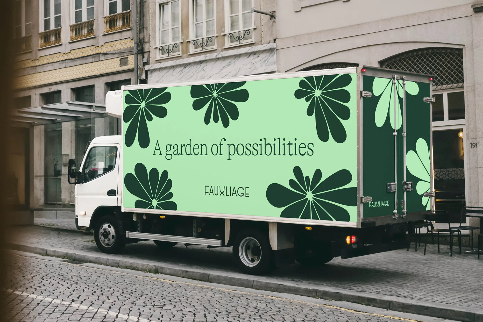 Delivery truck with floral graphics parked on a cobblestone street, featuring the text 'A garden of possibilities' and the brand name 'FAUXLIAGE'.