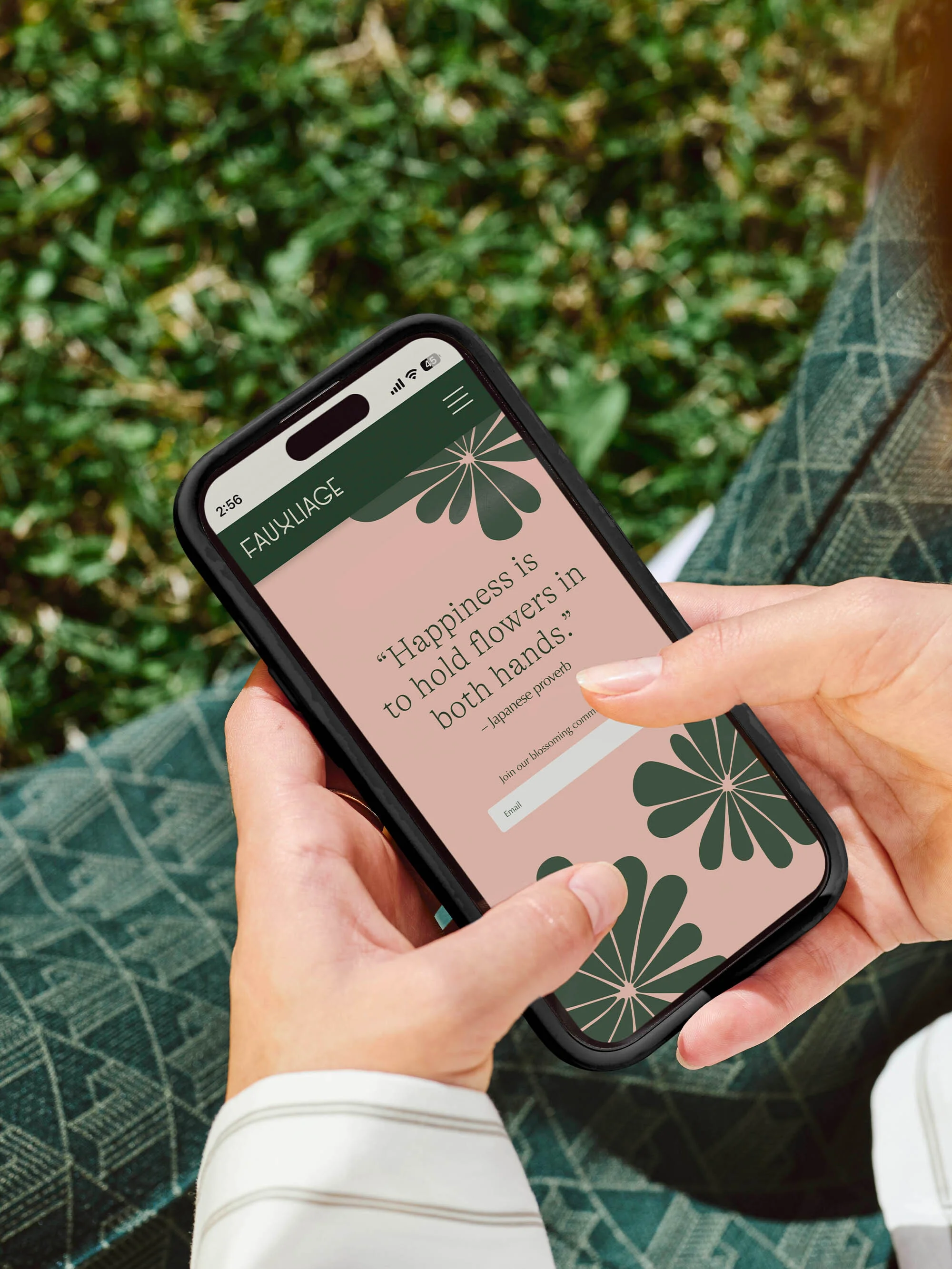 Close-up of a person holding a smartphone with a floral design website, set against a grassy background. Focus on the phone screen.