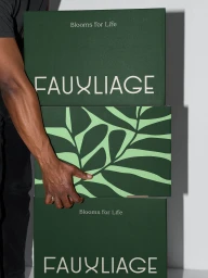 Man holding a stack of dark green branded Fauxliage boxes against a grey studio backdrop.