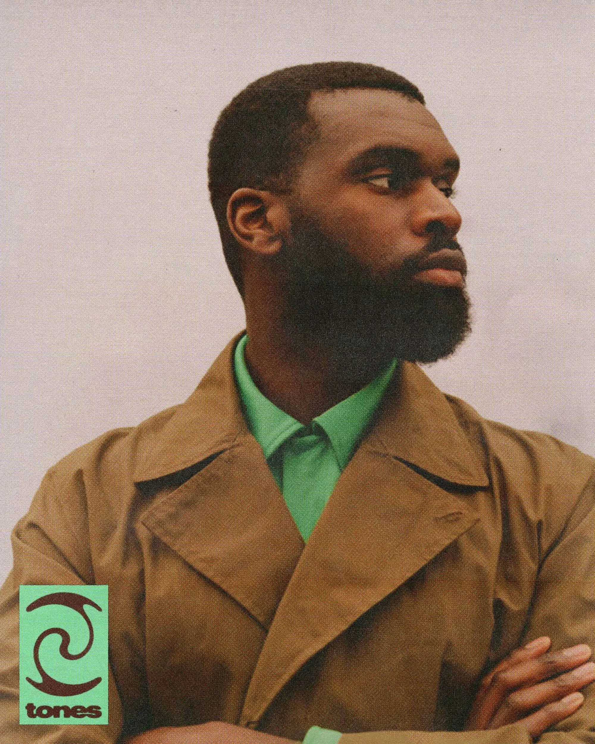 Close-up portrait of a Black man wearing a brown coat and green shirt, arms crossed, looking to the right, against a light background.