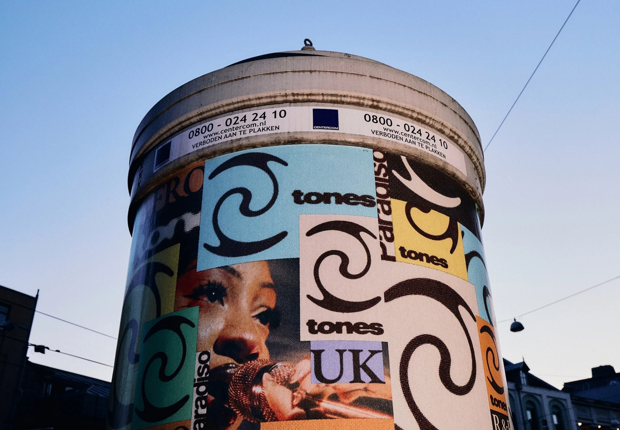 Cylindrical advertising column covered in music posters and contact info, set against a pale blue sky in an outdoor urban setting.