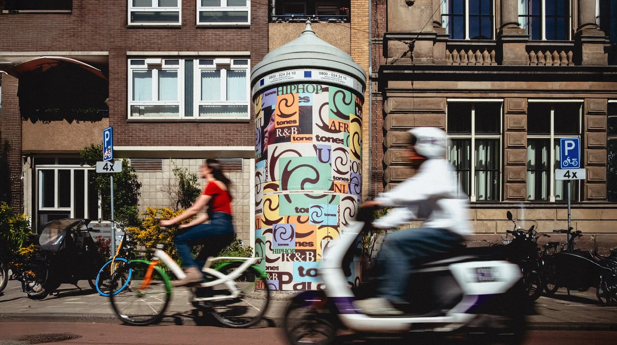 Woman riding bicycle, man on moped pass column with posters in European city. Bikes parked against building in background.