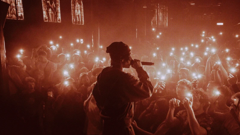 Singer performing on stage in orange lighting with a large crowd holding up their phones, capturing the performance in a concert venue.
