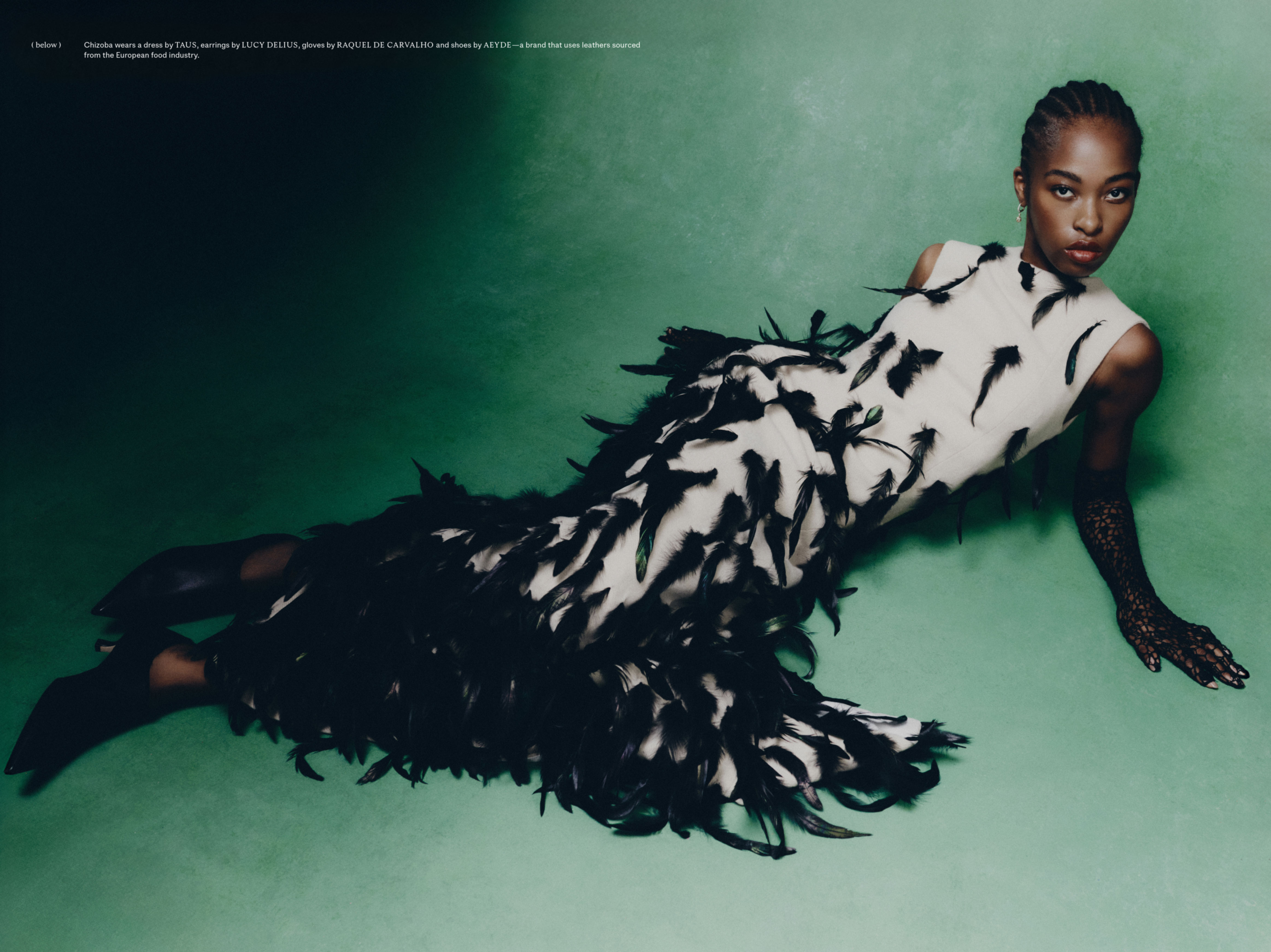 Woman in a cream and black feather dress, lying on a green surface, wearing black lace gloves, and black pointy-toe heeled shoes.