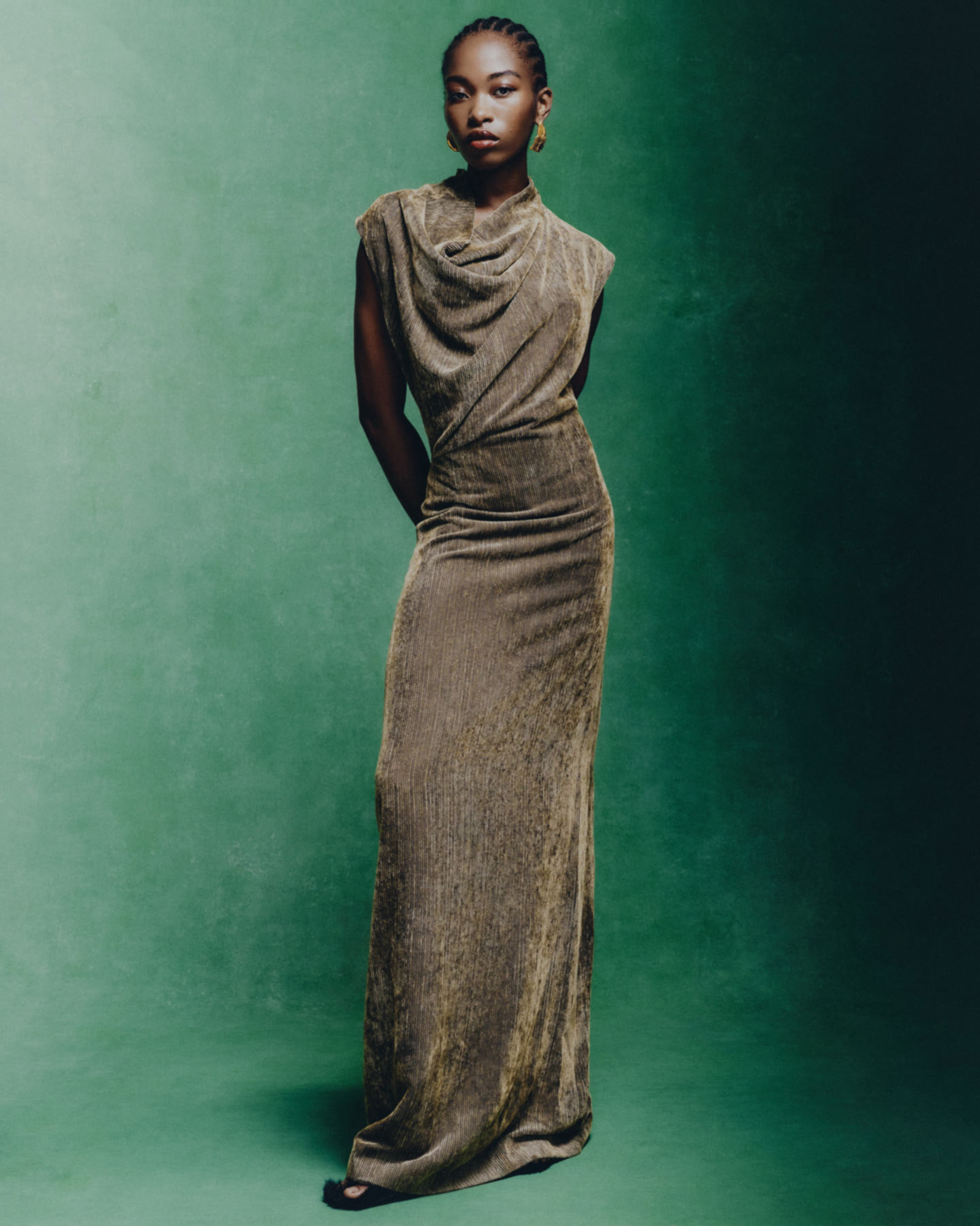Elegant African American woman in a draped golden gown poses against a green backdrop in a fashion editorial shot with natural light.