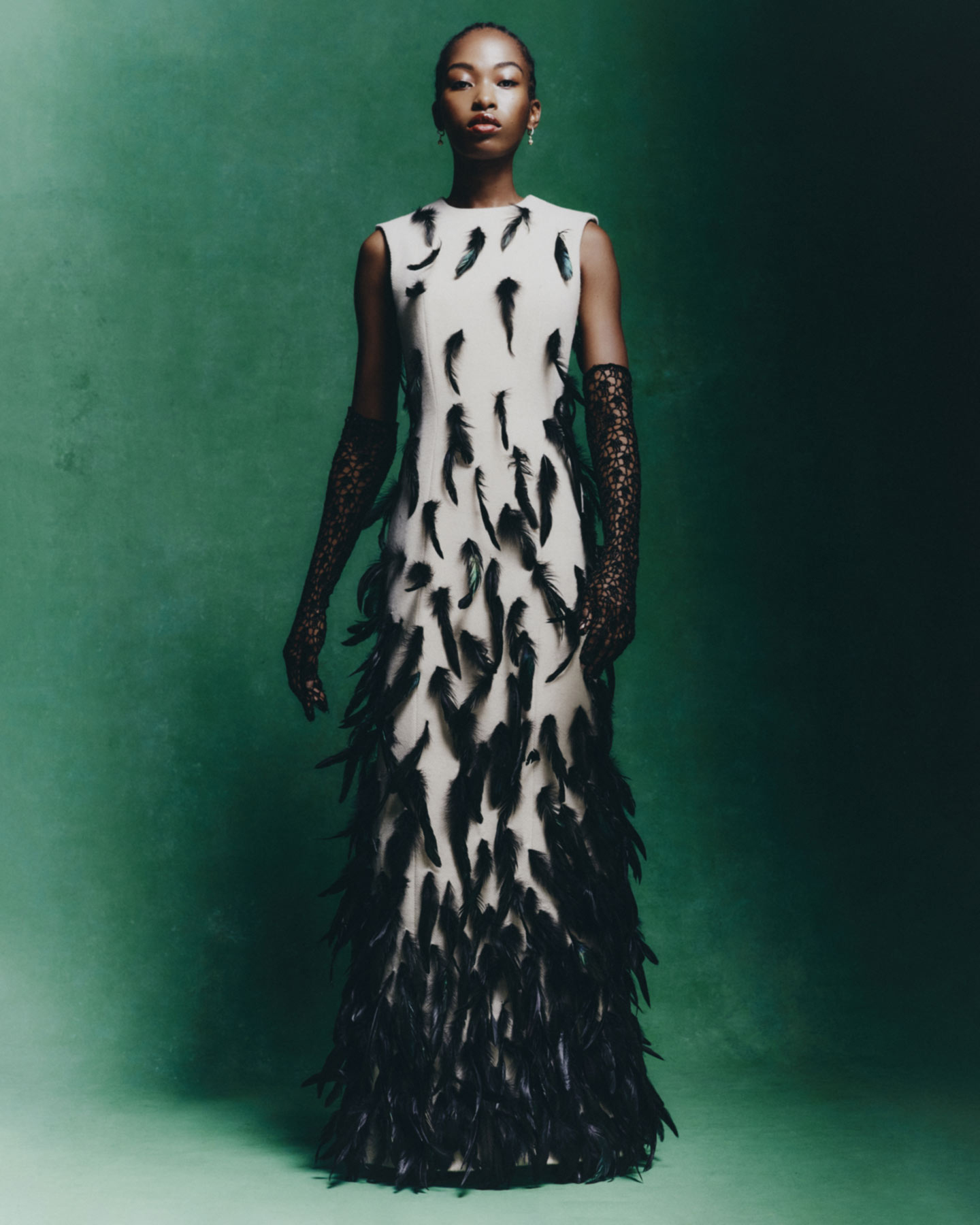 Elegant African American woman in an off-white gown with black feather detail, wearing lace gloves, posing against a green backdrop.
