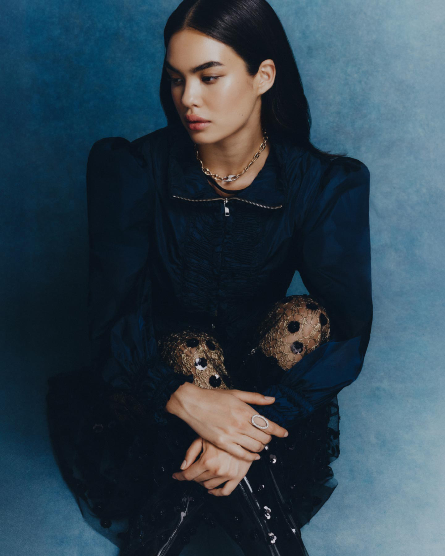 Portrait of a young woman with long dark hair wearing a navy top, gold necklace, and ring, sitting indoors against a textured blue backdrop.