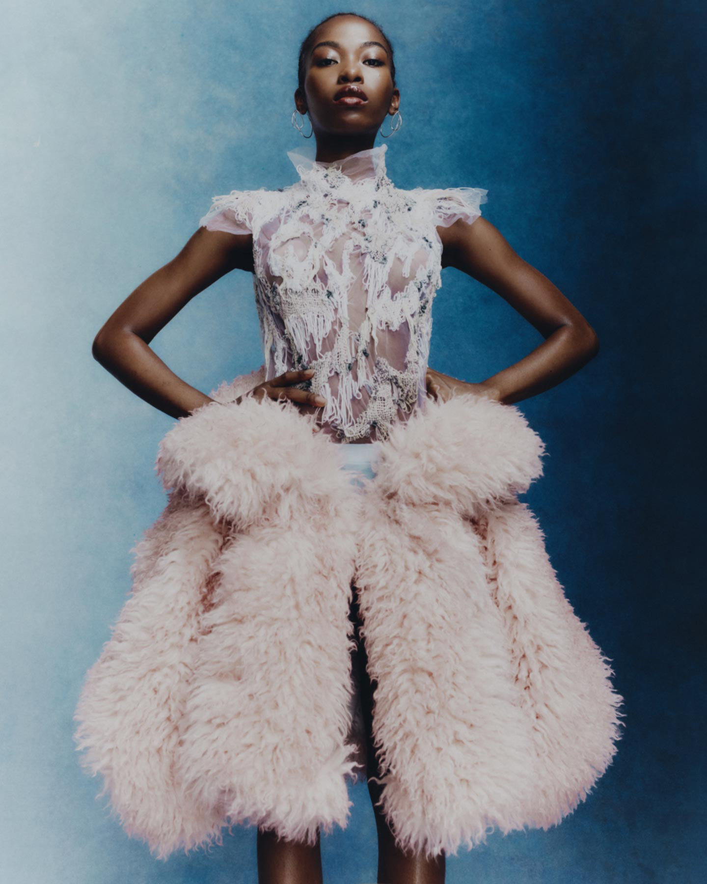 Portrait of Black female fashion model in lace dress and oversized fur coat, posing with hands on her hips against blue backdrop.