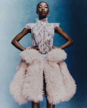 Portrait of Black female fashion model in lace dress and oversized fur coat, posing with hands on her hips against blue backdrop.