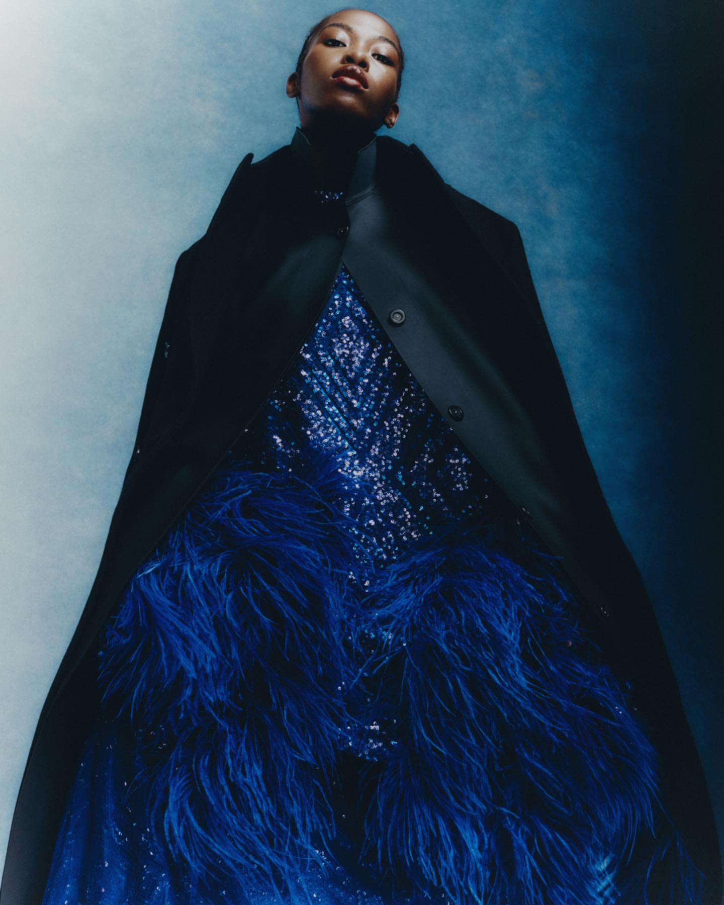 Elegant woman poses wearing a black coat over a blue sequined and feathered gown in a fashion editorial with cool studio lighting.