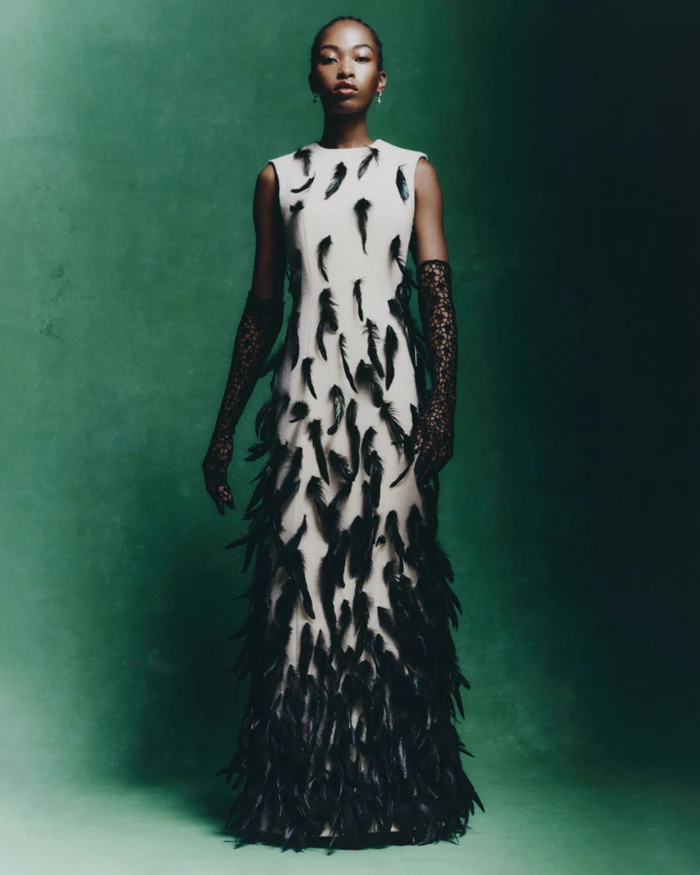 Model in sleeveless cream dress adorned with black feathers and lace gloves against a mottled green backdrop.