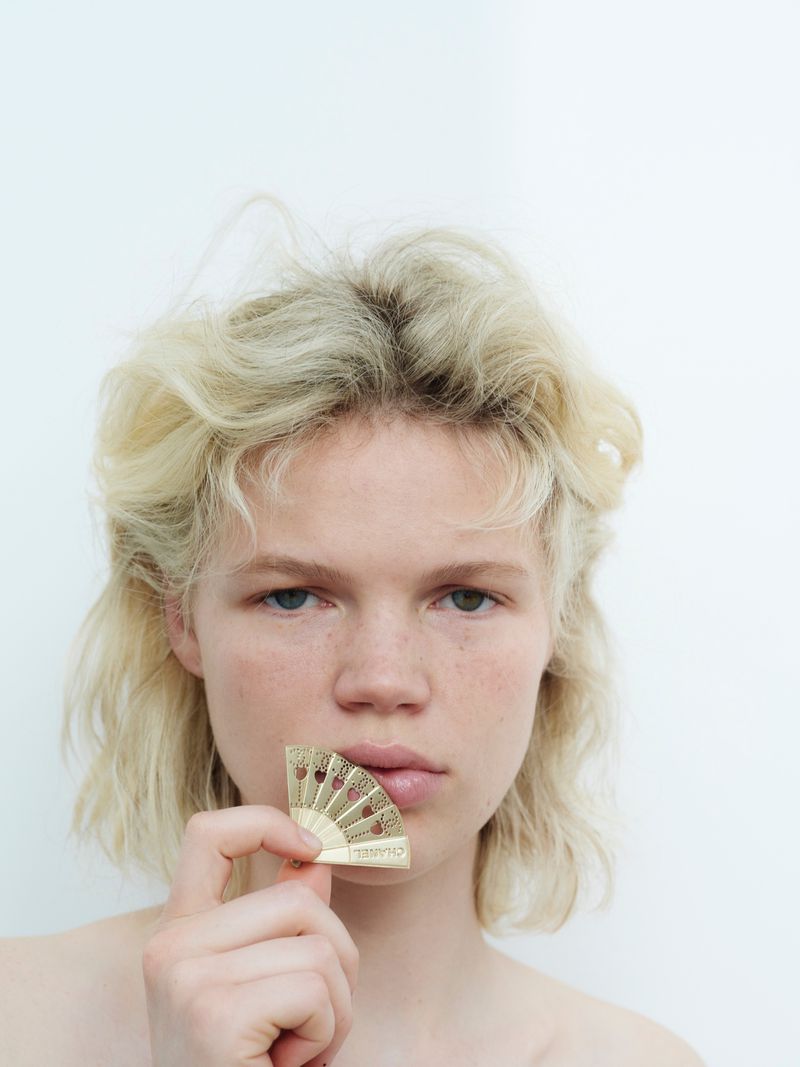 Blonde-haired woman holds miniature fan to her lips in a bright, high-key beauty shot against a white background indoors.