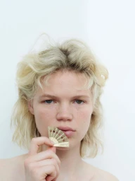 Close-up portrait of a blonde model holding a small decorative gold folding fan near her lips against a white background.