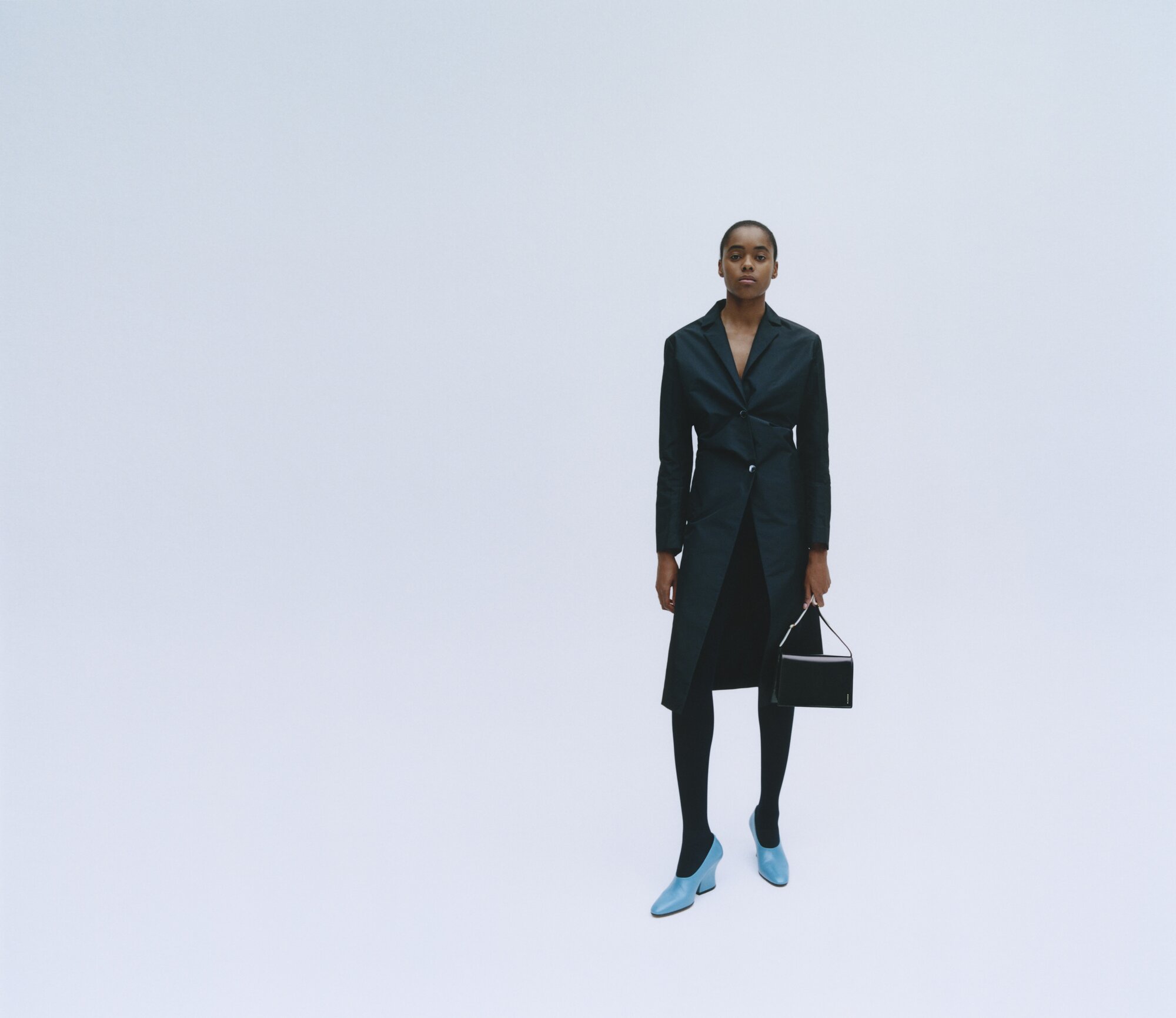 Fashionable woman poses in a black blazer dress, stockings, and light blue heels with a black purse in front of a plain white background.