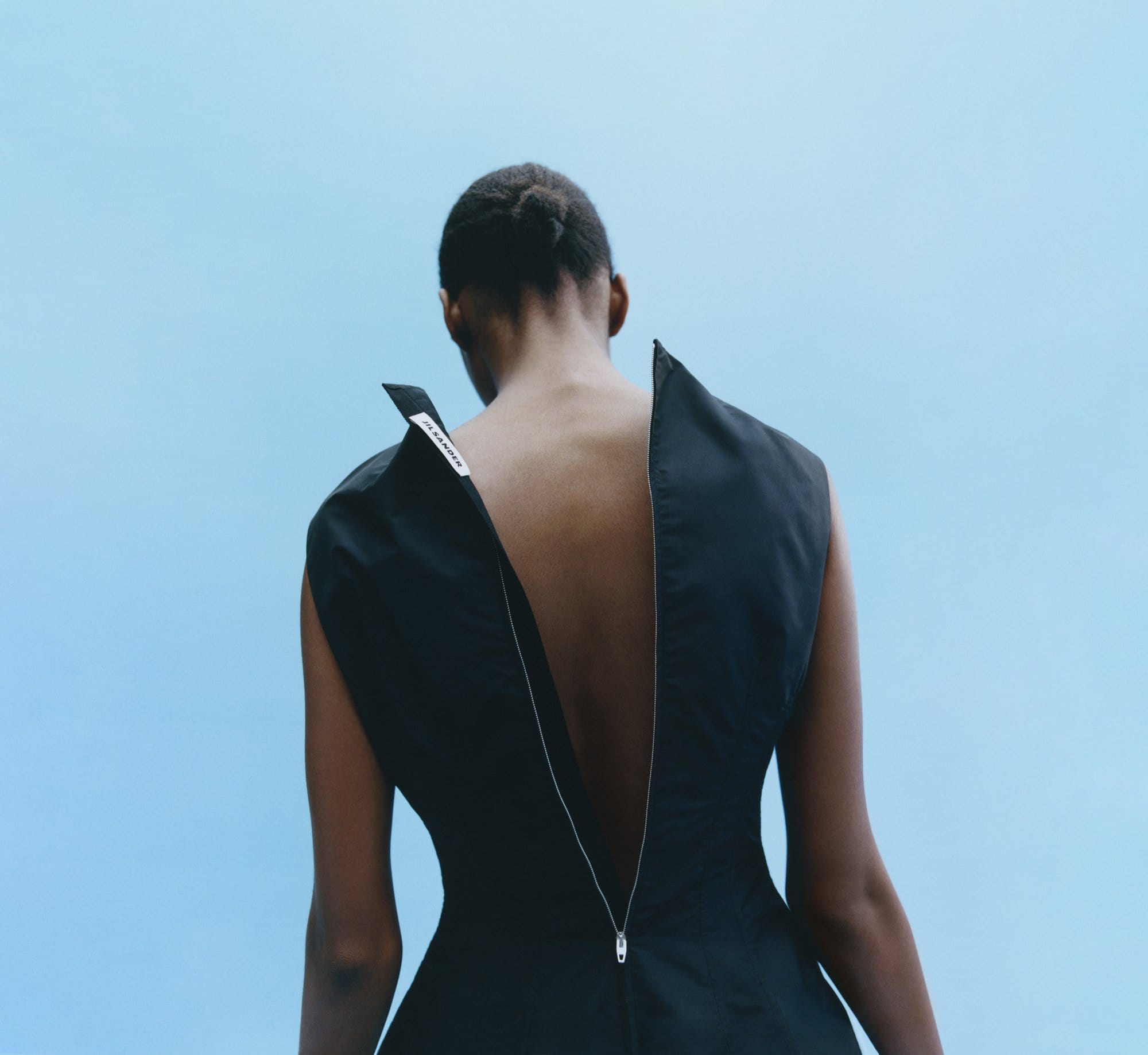 Rear view of Black fashion model wearing sleeveless black dress with unzipped back showing fair skin, against light blue sky background.