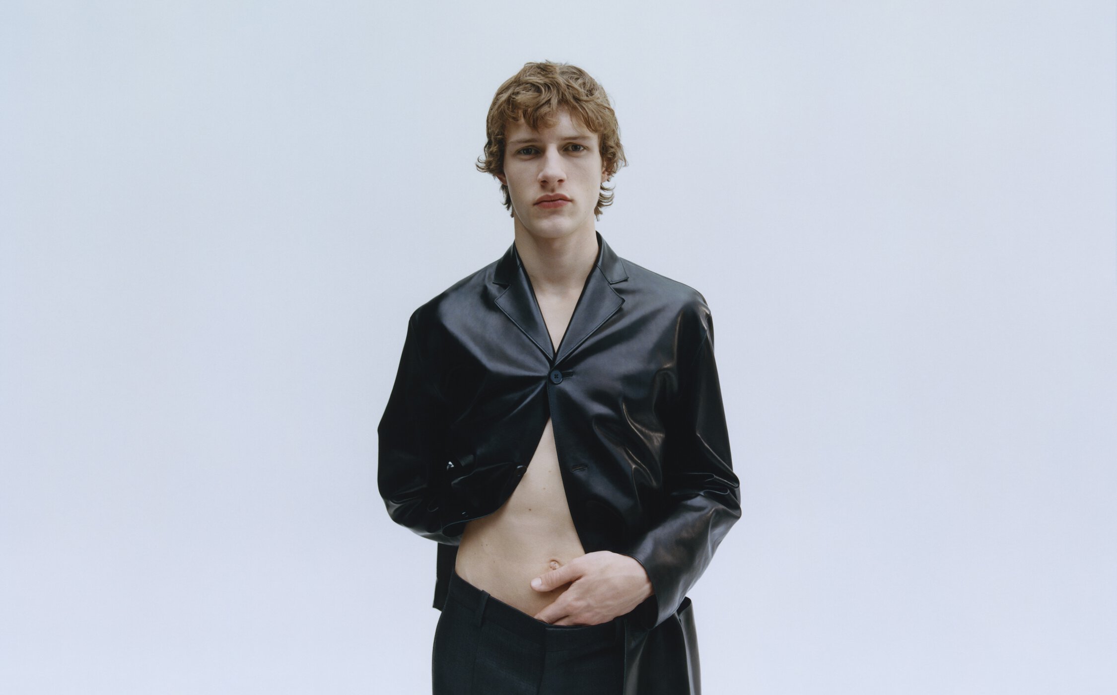 A young man with curly brown hair poses in a black blazer and pants against a white backdrop in a fashion forward studio portrait.